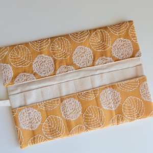 May include: A mustard-yellow fabric case with a white leaf and circle pattern. The case is open, revealing a beige interior. The fabric has a quilted texture. A white ribbon is attached to the side. The case is likely for storing items.