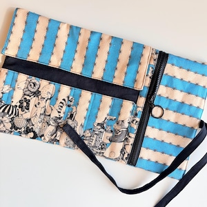 May include: A rectangular clutch with a blue and beige striped pattern. The clutch features a black zipper, a black strap, and a flap with a black border. The lower portion of the clutch has a black and white illustration.