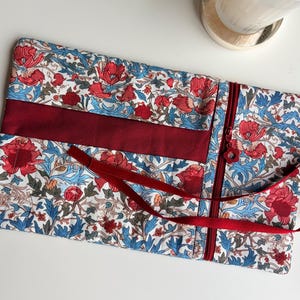 May include: A floral patterned case with a red zipper and a burgundy ribbon. The case features a white background with red flowers and blue leaves. A burgundy ribbon is tied around the case.