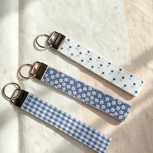 May include: Three fabric wristlet keychains with metal hardware and split rings. One features a white background with blue polka dots, another has a blue background with white daisy flowers, and the third has a blue and white gingham check pattern.
