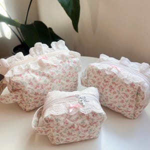 May include: Three floral print cosmetic bags with white lace trim and pink bows. The bags are in varying sizes, with a quilted pattern and zip closures. The fabric features a pattern of pink and blue flowers on a white background.