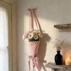 May include: A pink and white gingham fabric flower holder with a bow at the top and bottom, filled with daisies and pink flowers. The holder is suspended from the wall by a matching ribbon. A charming floral home decor item.