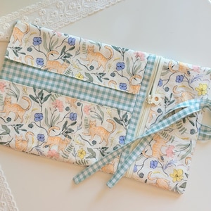 May include: A fabric roll-up organizer featuring a pattern of orange cats, green leaves, and small blue and yellow flowers. The case includes a light green and white gingham check fabric panel, a white zipper closure, and matching gingham fabric ties.