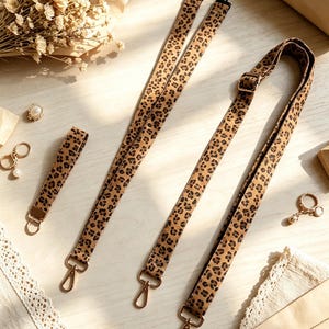 Peut inclure: Un ensemble de trois sangles en tissu à motif léopard avec des mousquetons pivotants en métal doré et une boucle de réglage sur la sangle la plus longue. Les sangles sont en tissu beige avec un motif animalier noir et marron foncé.