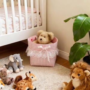 Puede incluir: Una colección de juguetes de animales de peluche, incluyendo un león, un elefante y una jirafa, están colocados sobre una alfombra color crema. Un osito de peluche está en una cesta rosa de cuadros con encaje. Una cuna blanca está en el fondo.
