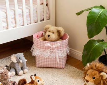 May include: A collection of plush animal toys, including a lion, elephant, and giraffe, are arranged on a cream-colored rug. A teddy bear sits in a pink gingham basket trimmed with lace. A white crib is in the background.