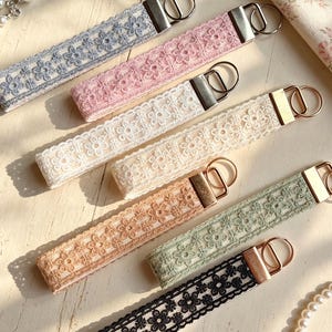 May include: A collection of six rectangular fabric key fobs featuring intricate floral lace patterns in blue, pink, cream, beige, sage green, and black. Each strap is secured with a silver-toned metal clamp and a circular split ring for keys.