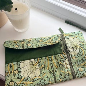 May include: A green and floral patterned clutch with a zipper closure. The clutch features a flap and a dark green accent panel. The floral pattern includes white flowers and green leaves. A lit candle in a glass is in the background.