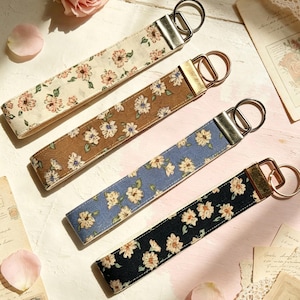 May include: Four fabric wristlet keychains featuring floral patterns in cream, brown, blue, and black. Each keychain has a silver-toned metal clasp and a circular split ring attached to one end, showcasing a repeating design of small light-coloured flowers.