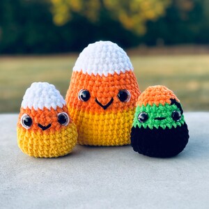 May include: Three plush crochet toys featuring two candy corn shapes with white, orange, and yellow yarn, and one smaller rounded toy with orange, green, and black yarn. Each toy has black plastic safety eyes and a stitched black mouth.