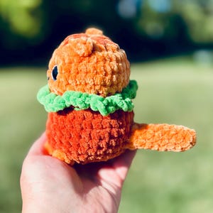 May include: A handmade orange crochet creature with a green collar and a small tail. The plush toy has a round head and body with small black eyes. The toy is held in a hand against a blurred green background.