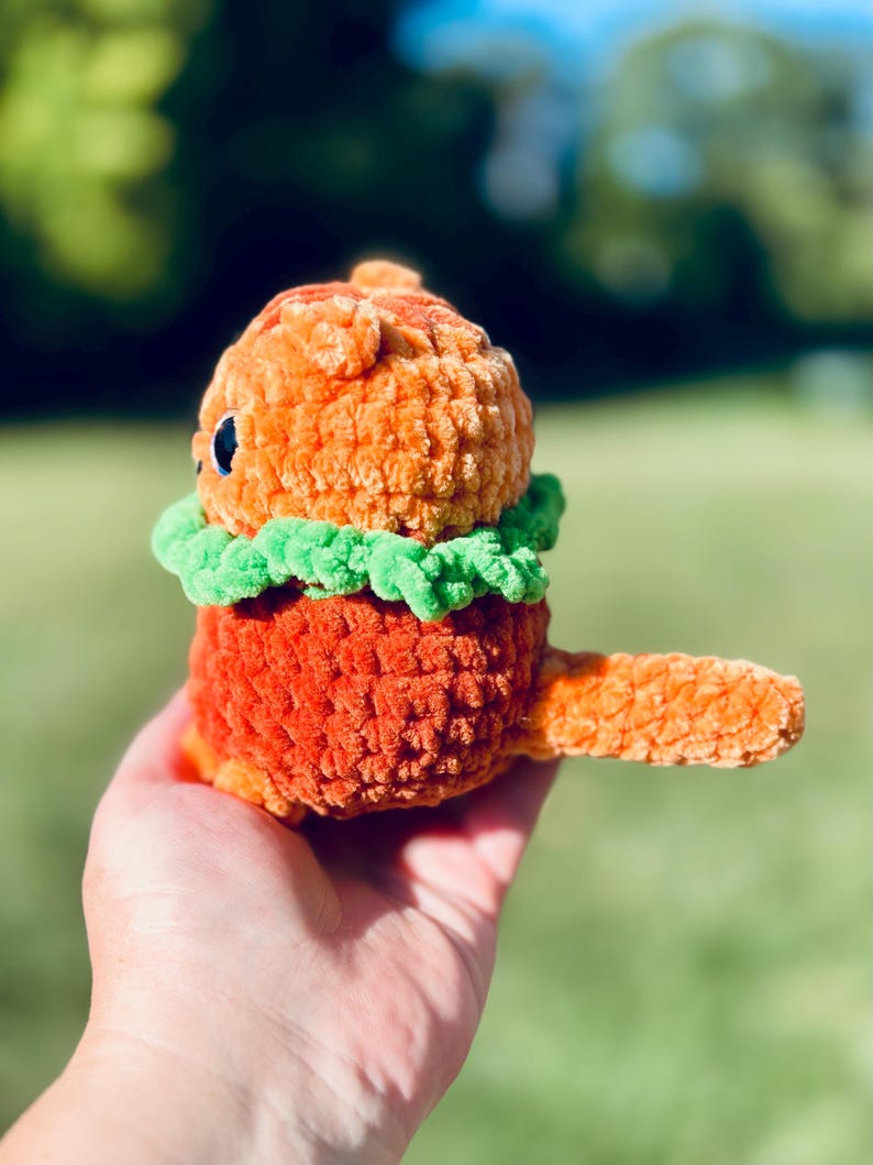 May include: A handmade orange crochet creature with a green collar and a small tail. The plush toy has a round head and body with small black eyes. The toy is held in a hand against a blurred green background.