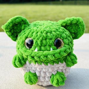 May include: A plush crochet creature featuring a vibrant green upper body and a light beige belly. The toy has large, round, glittery gold and black eyes, two small white fangs protruding from the mouth, and two prominent, rounded ears on top.