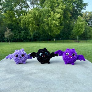 May include: Three small, round, crocheted plush bats arranged in a row. The left bat is light purple, the center bat is black with subtle sparkles, and the right bat is vibrant purple. Each bat features large black eyes and two small white fangs.