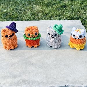 Crochet cats in a line that include an orange witch cat with a purple witch hat, orange pumpkin cat with green collar, gray cat with a green witch hat, and a candy corn cat with white, orange, and yellow yarn colors.