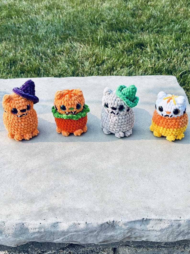 Crochet cats in a line that include an orange witch cat with a purple witch hat, orange pumpkin cat with green collar, gray cat with a green witch hat, and a candy corn cat with white, orange, and yellow yarn colors.
