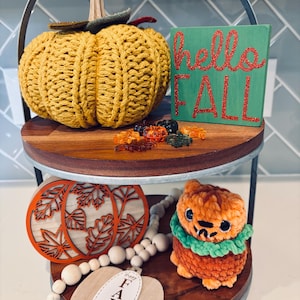May include: A two-tiered decorative tray with fall-themed items. The top tier features a knitted yellow pumpkin, a green sign with "hello FALL" in red glitter, and decorative fall-colored leaves. The bottom tier has wooden pumpkin cutouts, a pumpkin-shaped plush, and a beaded garland.