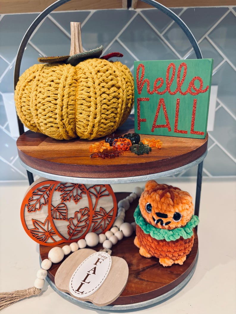 May include: A two-tiered decorative tray with fall-themed items. The top tier features a knitted yellow pumpkin, a green sign with "hello FALL" in red glitter, and decorative fall-colored leaves. The bottom tier has wooden pumpkin cutouts, a pumpkin-shaped plush, and a beaded garland.