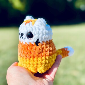 May include: A handmade crochet cat in the shape of candy corn. The cat has a white head with orange accents, large black eyes, and a tail with a white tip. The body is orange and yellow.