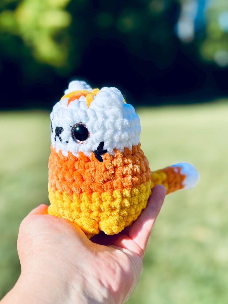 May include: A handmade crochet cat in the shape of candy corn. The cat has a white head with orange accents, large black eyes, and a tail with a white tip. The body is orange and yellow.