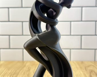 May include: A matte black sculpture depicting two abstract figures in an embrace. The figures are intertwined with smooth, flowing lines, creating a continuous form that curves upward. The surface shows a subtle 3D printed texture throughout.