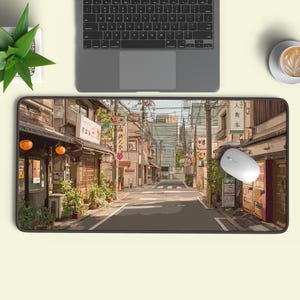 May include: A rectangular mouse mat with a detailed illustration of a Japanese street scene. The image includes buildings, streetlights, and power lines. A laptop, a white mouse, and a cup of coffee are also visible.