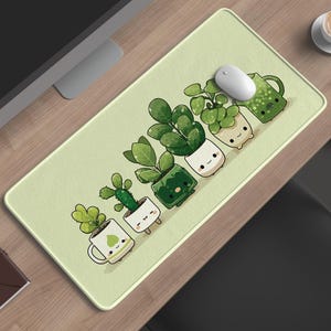 Puede incluir: Alfombrilla de ratón rectangular verde claro con una fila de plantas en macetas de dibujos animados con caras sonrientes. Un ratón de ordenador blanco está sobre la alfombrilla. También se ven una taza de café y un pequeño cuaderno marrón.