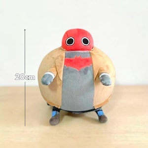 May include: A plush toy of a character with a red helmet and large black eyes. The figure is wearing a tan jacket over a gray shirt with a red emblem. The toy is approximately 8 inches tall.