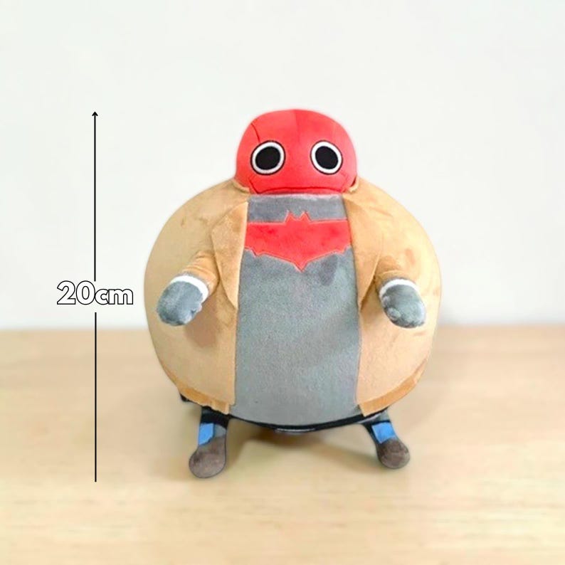 May include: A plush toy of a character with a red helmet and large black eyes. The figure is wearing a tan jacket over a gray shirt with a red emblem. The toy is approximately 8 inches tall.