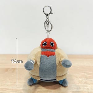 May include: A plush keychain featuring a round, robot-like character. The toy has a red head with black eyes, a gray torso, and a tan jacket. The keychain is approximately 4.7 inches (12 cm) tall and has a silver metal clasp.
