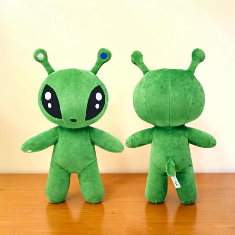 May include: Two green plush alien toys with large black eyes, small arms, and antennae. One alien has a blue dot on its antenna. The toys are standing on a wooden surface.