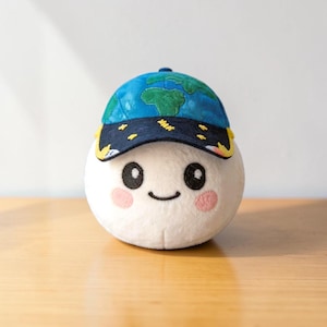 May include: A plush toy with a white body, black eyes, and a smiling mouth. It wears a blue cap with a green map design and yellow accents. The toy has pink cheeks and is set on a wooden surface.