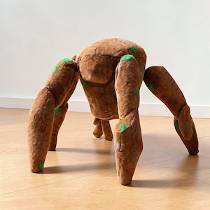 Peut inclure: Un tabouret en forme d'araignée en peluche marron à six pattes. Le tabouret a un corps arrondi et des pattes avec des accents verts. Les pattes sont positionnées pour supporter le poids du tabouret, créant un meuble unique et fantaisiste.