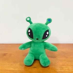 May include: A green plush alien toy with large black eyes and small antennae. The alien has a small nose and is sitting with its arms and legs outstretched. The antennae have white and blue accents. The toy is made of soft material.