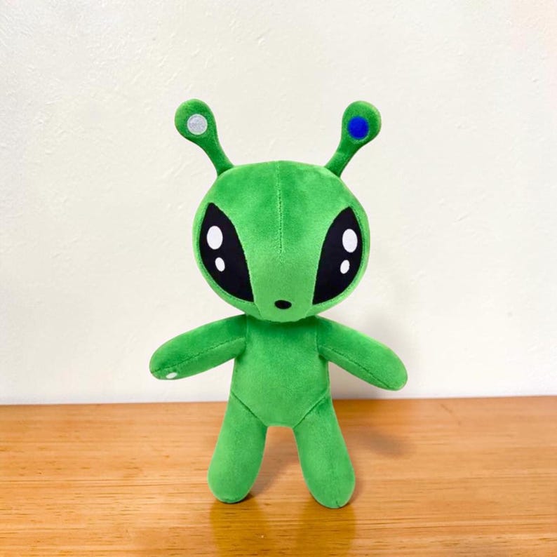 May include: A plush green alien toy with large black eyes and small arms and legs. The alien has two antennae, one with a silver dot and the other with a blue dot. The toy is standing on a wooden surface.