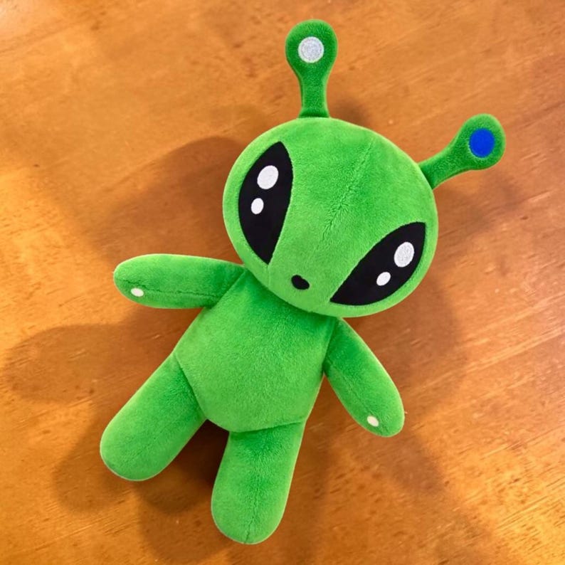 May include: A plush green alien toy with large black eyes and white highlights. The alien has two antennae with white and blue accents, and white spots on its hands and feet. The toy is lying on a wooden surface.