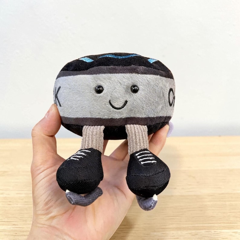 Pode incluir: Um brinquedo de pel&uacute;cia em forma de fita cassete com um rosto sorridente. O brinquedo &eacute; principalmente cinza e preto, com sapatos pretos e meias bege. O brinquedo tem a letra "K" de um lado e o n&uacute;mero "0" do outro.