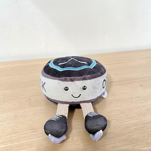 Pode incluir: Um brinquedo de pel&uacute;cia em forma de disco de h&oacute;quei com um rosto sorridente, sapatos pretos e pernas bege. O disco &eacute; preto com um desenho turquesa e branco no topo e a letra "K" na lateral. O brinquedo est&aacute; sobre uma superf&iacute;cie de madeira.