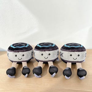 Pode incluir: Tr&ecirc;s brinquedos de pel&uacute;cia em forma de personagens redondos e sorridentes, com detalhes em preto e cinza. Cada um tem um topo preto com uma faixa azul, um corpo branco com "OK" impresso e p&eacute;s pretos. Os brinquedos est&atilde;o dispostos em fila.