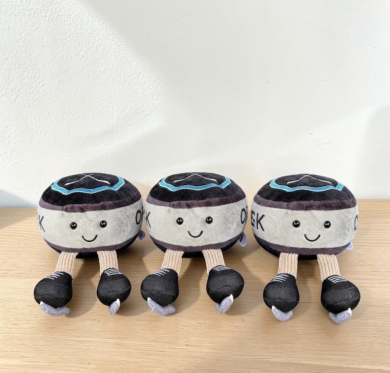 Pode incluir: Tr&ecirc;s brinquedos de pel&uacute;cia em forma de personagens redondos e sorridentes, com detalhes em preto e cinza. Cada um tem um topo preto com uma faixa azul, um corpo branco com "OK" impresso e p&eacute;s pretos. Os brinquedos est&atilde;o dispostos em fila.