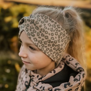 Könnte beinhalten: Ein Stirnband mit tan-schwarzem Leopardenmuster. Die Person trägt eine passende, gepolsterte Jacke mit Leopardenmuster in Tan und Schwarz. Die Jacke hat eine schwarze Kapuze und einen Reißverschluss.