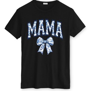May include: Black t-shirt featuring the word "MAMA" in a blue and white floral design, arched across the chest. A matching floral bow is positioned below the word. The shirt has a crew neck and short sleeves.
