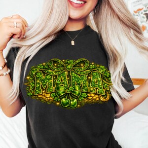 Peut inclure: T-shirt noir avec un motif de la Saint-Patrick. Le mot "MAMA" est écrit en trèfles verts, avec un nœud vert et des accents dorés. Des pièces d'or et un fer à cheval sont au bas du motif.