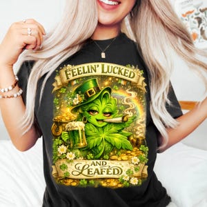 Peut inclure: T-shirt noir avec un personnage de feuille de cannabis de dessin animé portant un chapeau vert, tenant une bière et un joint allumé. Le motif comprend un arc-en-ciel, des pièces d'or et le texte "Feelin' Lucked" et "and Leafed".