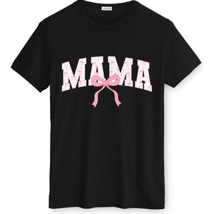 May include: Black crew neck t-shirt featuring the word "MAMA" in white, filled with pink bows. A large pink bow is positioned below the word. The shirt is a casual style.