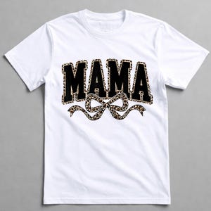 May include: White crew neck t-shirt with the word "MAMA" in black, leopard print filled letters. A leopard print bow is beneath the word. The shirt is made of a soft material.