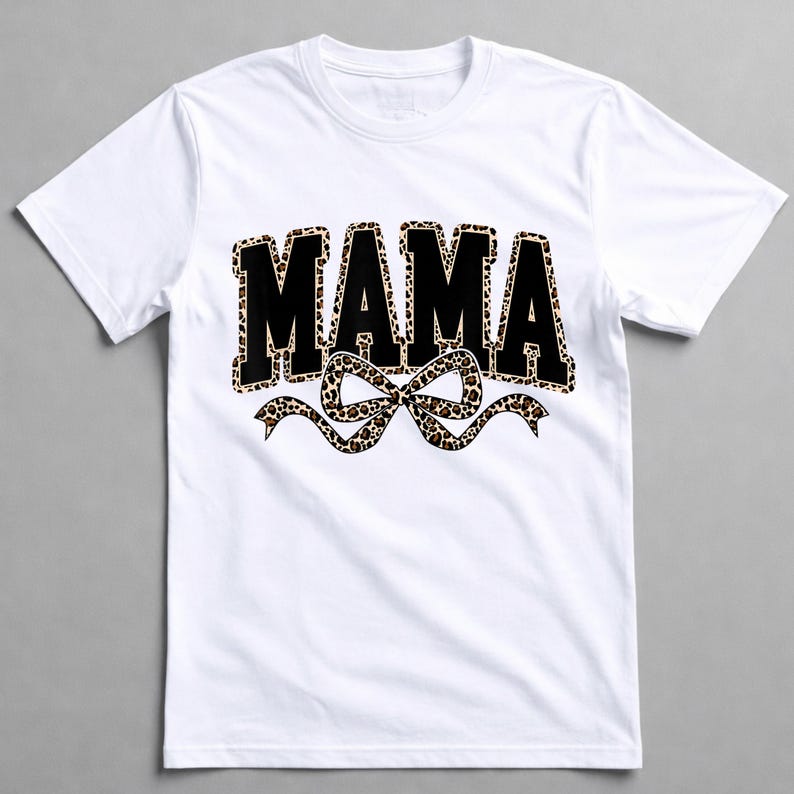 May include: White crew neck t-shirt with the word "MAMA" in black, leopard print filled letters. A leopard print bow is beneath the word. The shirt is made of a soft material.