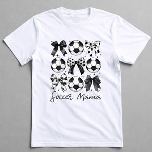 May include: White t-shirt featuring a design of soccer balls and bows in black and white. The words "Soccer Mama" are written in a script font below the graphic. The shirt has a crew neck and short sleeves.