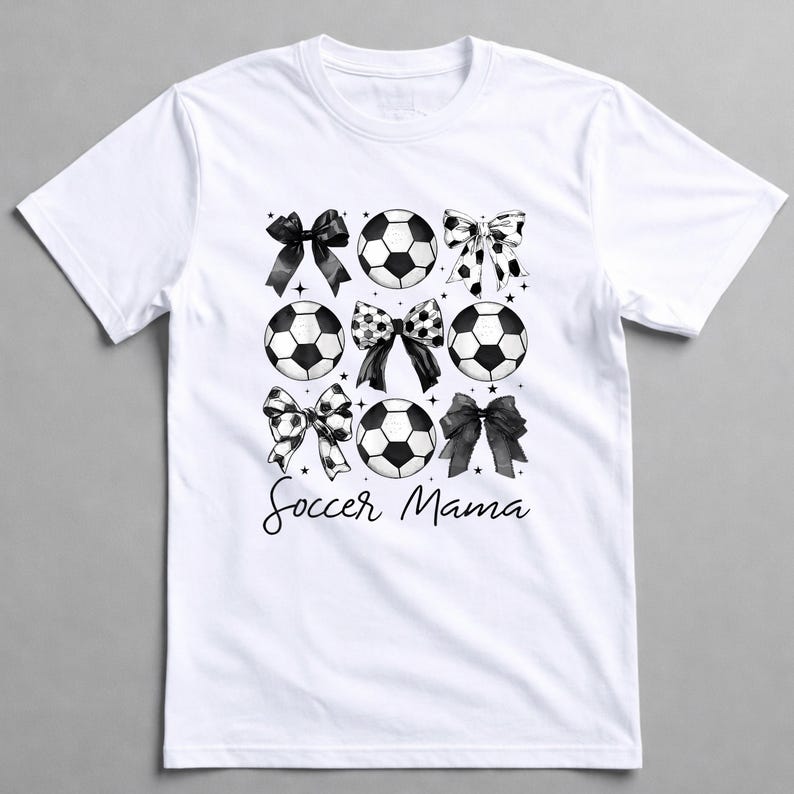 May include: White t-shirt featuring a design of soccer balls and bows in black and white. The words "Soccer Mama" are written in a script font below the graphic. The shirt has a crew neck and short sleeves.