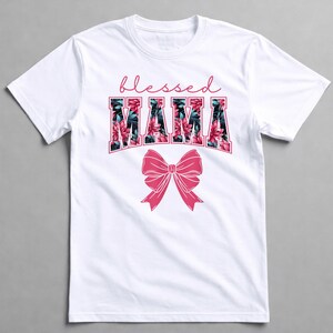 May include: White t-shirt featuring the text "blessed MAMA" in a floral design and a pink bow. The word "blessed" is in a cursive font above the word "MAMA". The floral design incorporates pink and green flowers.